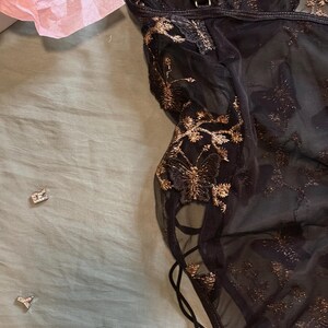 May include: Black lingerie with gold floral embroidery. The sheer fabric features intricate gold detailing and a delicate lace-up design on the side. The garment is partially visible, showcasing its elegant and detailed craftsmanship.