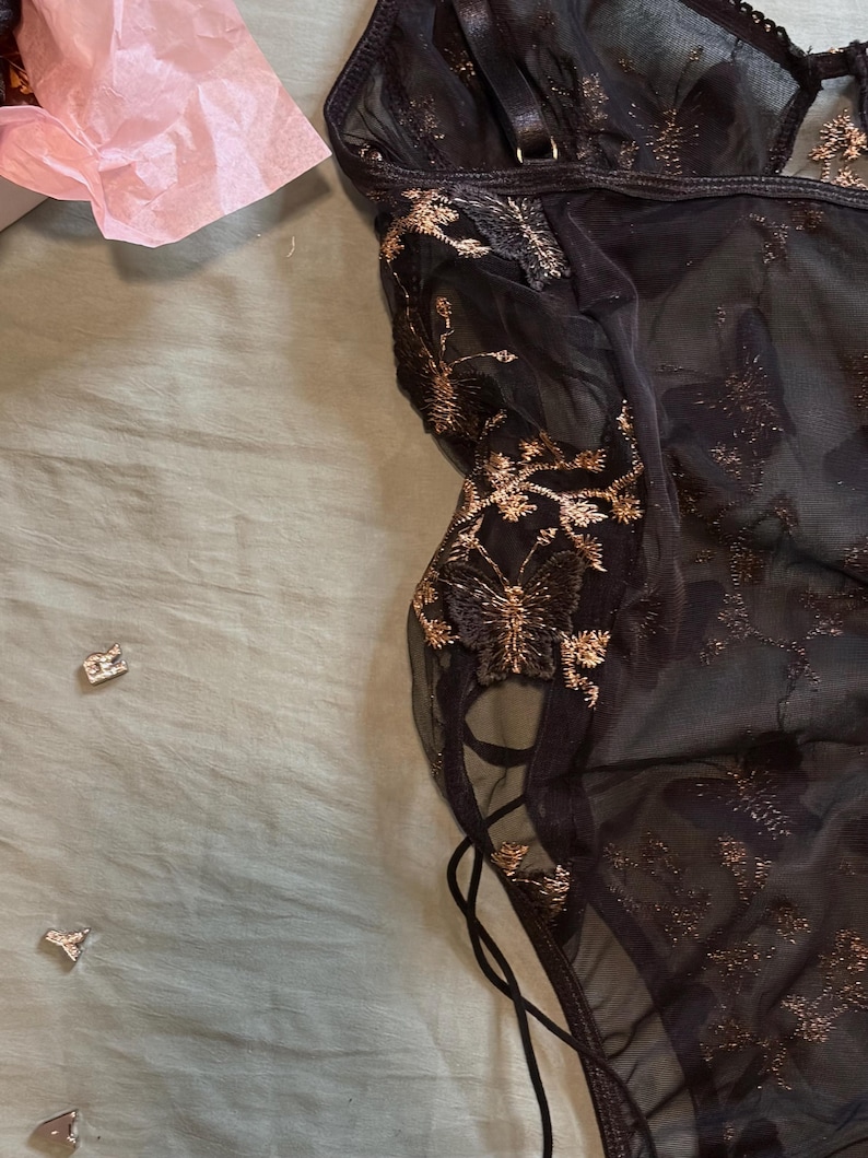 May include: Black lingerie with gold floral embroidery. The sheer fabric features intricate gold detailing and a delicate lace-up design on the side. The garment is partially visible, showcasing its elegant and detailed craftsmanship.