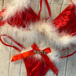 May include: Red lingerie set with white feather trim. The set includes a halter top and a thong, both with red satin fabric. A red ribbon bow on the thong features the word "NAME" in silver letters. The lingerie is designed with a festive theme.