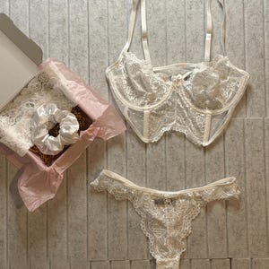 May include: A white lace lingerie set, including a bra and thong, displayed on a gray surface. A box with a scrunchie and lace trim is also present. The bra has adjustable straps and a delicate floral lace design.