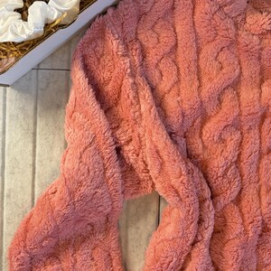May include: A coral-colored, cable-knit sweater with long sleeves. The sweater has a soft, fuzzy texture. A white satin scrunchie is visible in the upper left corner, inside a cardboard box.