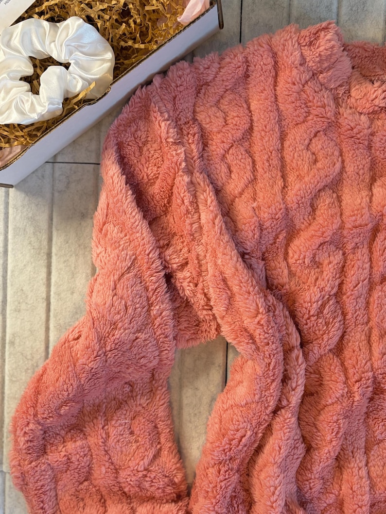 May include: A coral-colored, cable-knit sweater with long sleeves. The sweater has a soft, fuzzy texture. A white satin scrunchie is visible in the upper left corner, inside a cardboard box.