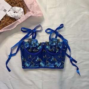 Puede incluir: Un bustier azul bordado con detalles florales. El bustier tiene correas de cinta azul y está decorado con flores azul claro y tallos verdes. Una scrunchie blanca y una caja rosa están en el fondo.