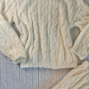 May include: Cream-colored, fuzzy, cable-knit sweater and matching bottoms. The soft, textured fabric creates a cozy look. The sweater has a high neckline and long sleeves. The bottoms are visible at the bottom of the image.