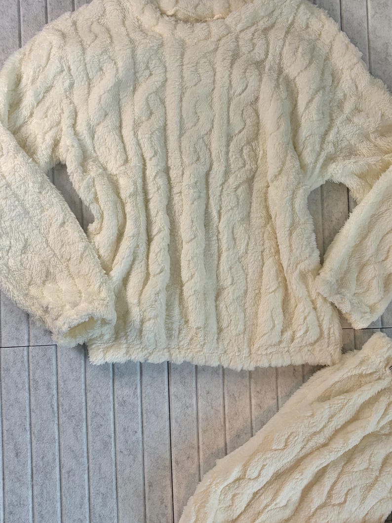 May include: Cream-colored, fuzzy, cable-knit sweater and matching bottoms. The soft, textured fabric creates a cozy look. The sweater has a high neckline and long sleeves. The bottoms are visible at the bottom of the image.
