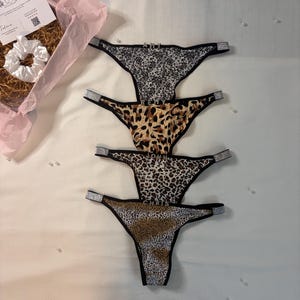 May include: Four thong underwear with animal print patterns, including leopard and cheetah prints. Each thong has a rhinestone accent on the sides. The underwear are displayed on a white surface.