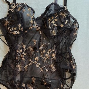 May include: Black lingerie bodysuit with gold floral embroidery. The sheer fabric features a delicate butterfly and flower design. The bodysuit has adjustable straps and lace-up detailing on the sides.