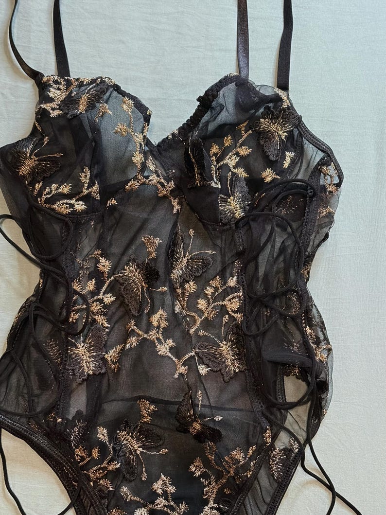 May include: Black lingerie bodysuit with gold floral embroidery. The sheer fabric features a delicate butterfly and flower design. The bodysuit has adjustable straps and lace-up detailing on the sides.