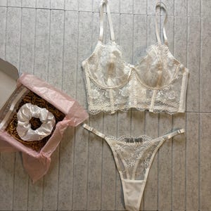May include: A white lingerie set featuring a bra, thong, and a scrunchie in a gift box. The bra and thong are adorned with delicate floral lace. The set is displayed on a gray tiled surface.