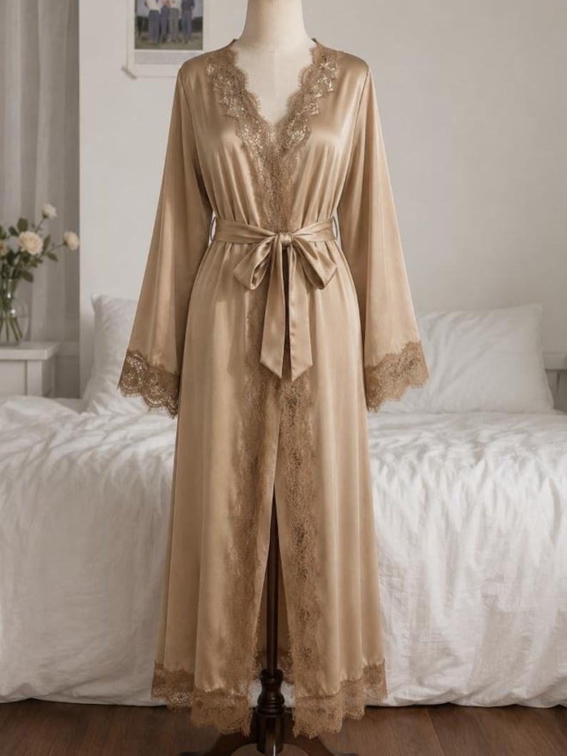 May include: A floor-length champagne-colored satin robe featuring long, wide sleeves and a V-neckline. The garment is adorned with intricate lace trim along the collar, sleeve cuffs, and hem, and includes a matching satin tie belt at the waist.