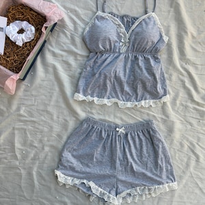 Puede incluir: Un conjunto de pijama de dos piezas en tela gris jaspeado que incluye una camiseta de tirantes finos, un escote en V adornado con encaje blanco y un pequeño lazo central. El pantalón corto a juego tiene una cintura elástica con un pequeño lazo y borde de encaje blanco.