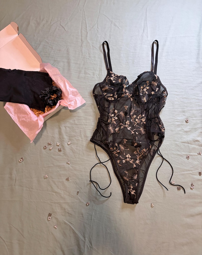 May include: Black lingerie bodysuit with floral embroidery and adjustable straps. The bodysuit is displayed on a light green surface next to a box with tissue paper and a black garment. Scattered letters are also visible.
