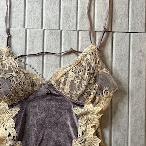 May include: A camisole featuring a dark mauve velvet bodice with cream-colored floral lace overlays on the cups and sides. The garment includes thin adjustable shoulder straps and a delicate string of small white faux pearls across the neckline.
