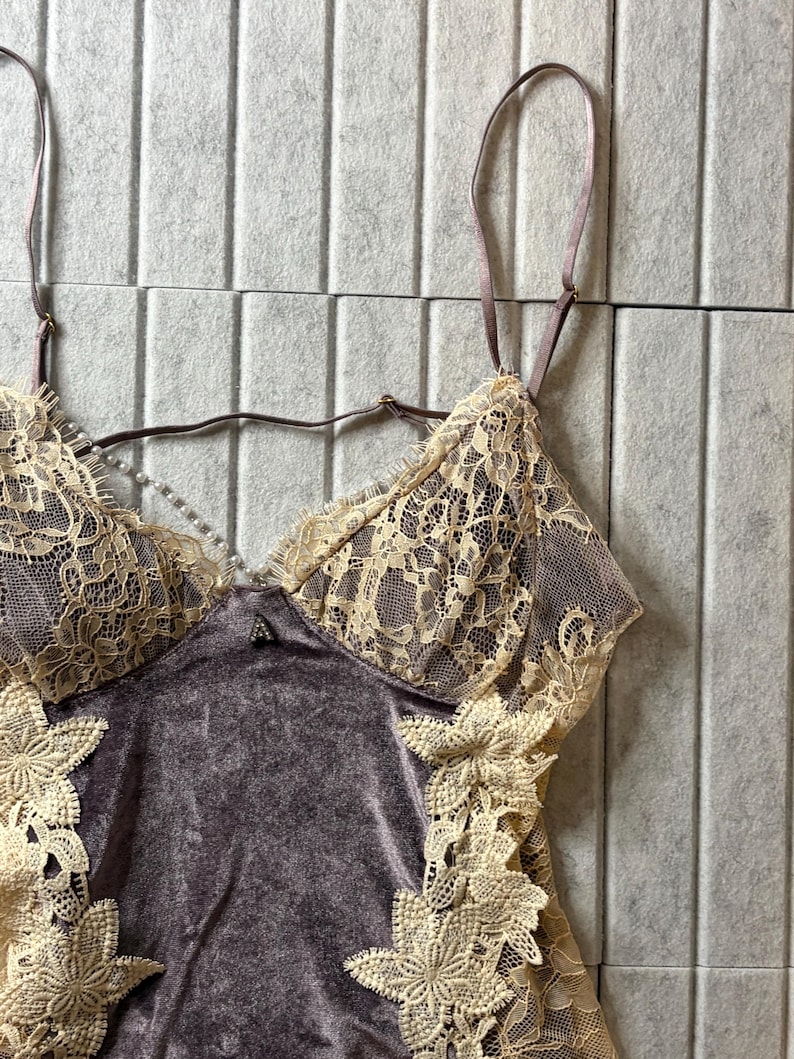 May include: A camisole featuring a dark mauve velvet bodice with cream-colored floral lace overlays on the cups and sides. The garment includes thin adjustable shoulder straps and a delicate string of small white faux pearls across the neckline.