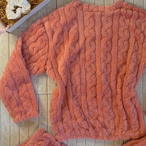 May include: A coral-colored, plush pajama set with a cable knit design. The set includes a long-sleeved top and matching bottoms. A white scrunchie and a gift box with tissue paper are also visible.