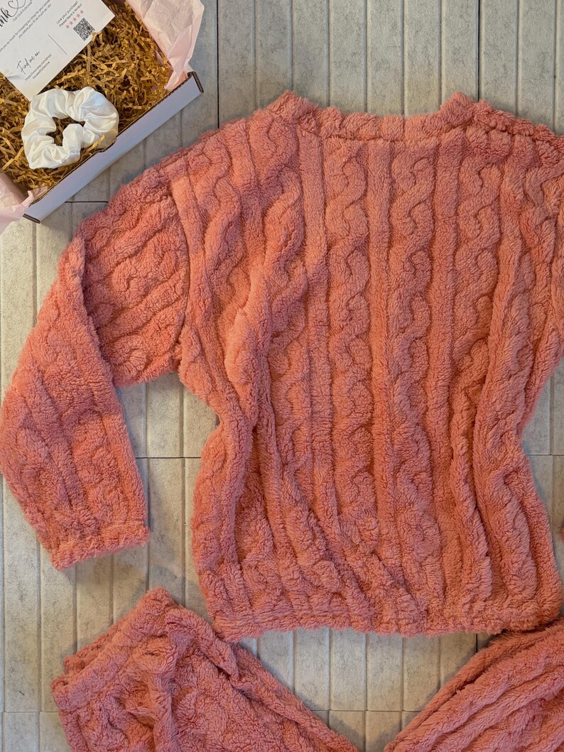 May include: A coral-colored, plush pajama set with a cable knit design. The set includes a long-sleeved top and matching bottoms. A white scrunchie and a gift box with tissue paper are also visible.