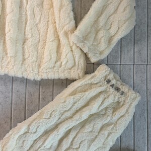 May include: Cream-colored, textured loungewear set. The set includes a long-sleeved top and matching pants. The pants' waistband features the word "MAME" in small, sparkling letters. The fabric has a soft, plush appearance.