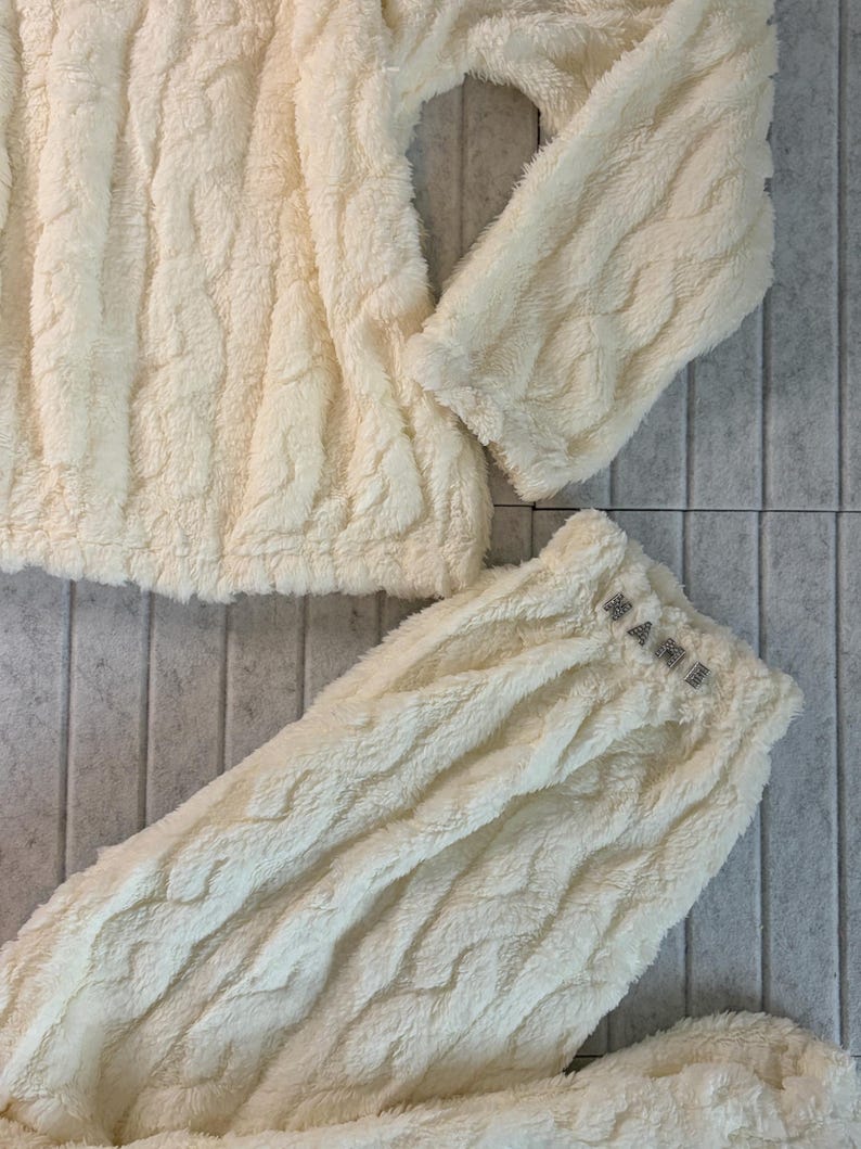 May include: Cream-colored, textured loungewear set. The set includes a long-sleeved top and matching pants. The pants' waistband features the word "MAME" in small, sparkling letters. The fabric has a soft, plush appearance.