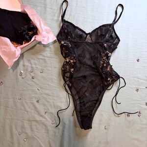May include: Black lingerie set with floral embroidery. The set includes a bodysuit with adjustable straps and side ties, and a matching item in a box with pink tissue paper. Scattered letters are on a light green surface.