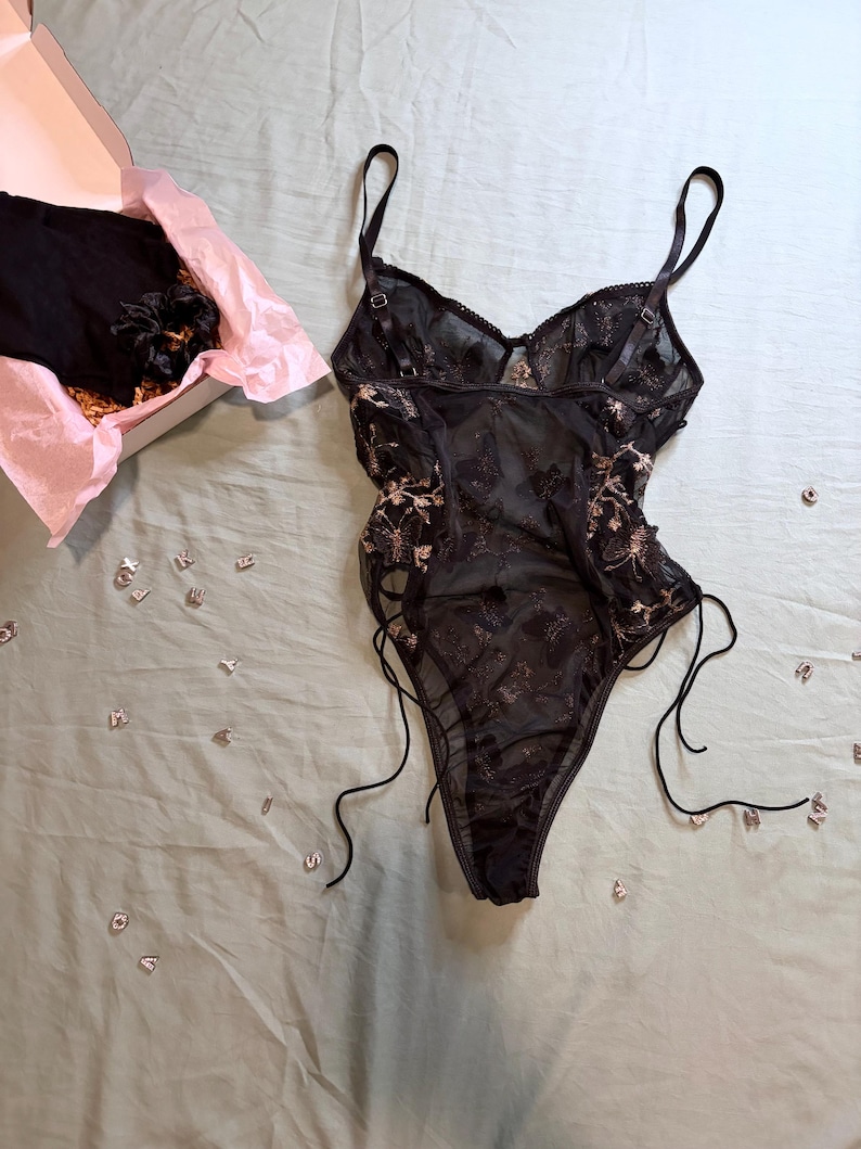 May include: Black lingerie set with floral embroidery. The set includes a bodysuit with adjustable straps and side ties, and a matching item in a box with pink tissue paper. Scattered letters are on a light green surface.