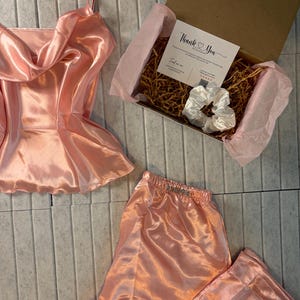 Op de afbeelding: Een perzikkleurige satijnen pyjamaset, bestaande uit een camisole top en broek, wordt gepresenteerd met een geschenkdoos. De doos bevat een witte scrunchie en een kaart met de tekst "Thank You". De set is gemaakt van een glanzende, gladde stof.