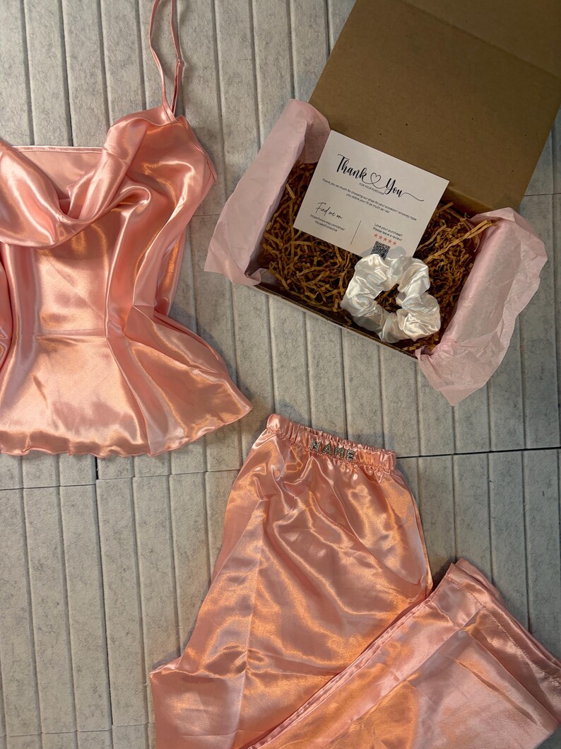 Op de afbeelding: Een perzikkleurige satijnen pyjamaset, bestaande uit een camisole top en broek, wordt gepresenteerd met een geschenkdoos. De doos bevat een witte scrunchie en een kaart met de tekst "Thank You". De set is gemaakt van een glanzende, gladde stof.