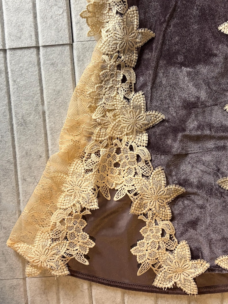 May include: A dark brown velvet garment featuring an intricate cream-colored floral lace trim along the edge. The lace displays a detailed petal pattern, contrasting with the smooth, soft texture of the dark brown fabric base underneath.