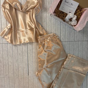 Op de afbeelding: Een champagnekleurige satijnen pyjamaset, bestaande uit een camisole top en een broek, wordt gepresenteerd op een lichtgrijze betegelde ondergrond. Een kleine geschenkdoos met een "Thank You" kaart en een witte scrunchie is ook aanwezig. De set heeft een glanzende, luxueuze uitstraling.