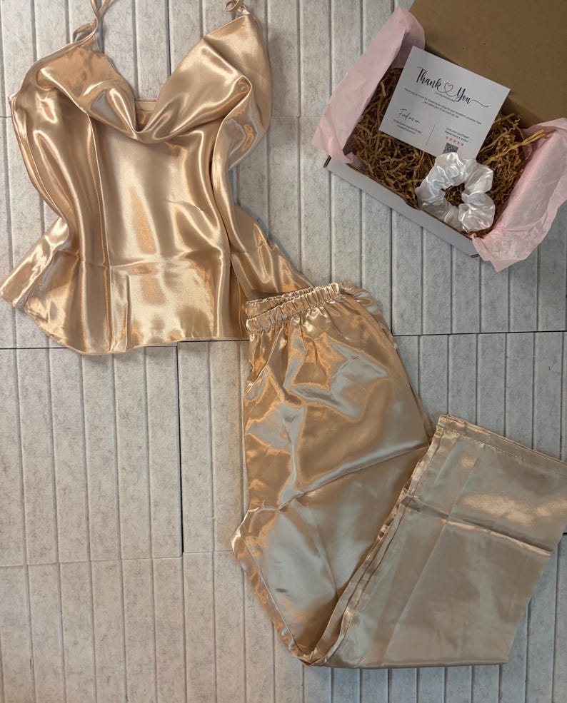 Op de afbeelding: Een champagnekleurige satijnen pyjamaset, bestaande uit een camisole top en een broek, wordt gepresenteerd op een lichtgrijze betegelde ondergrond. Een kleine geschenkdoos met een "Thank You" kaart en een witte scrunchie is ook aanwezig. De set heeft een glanzende, luxueuze uitstraling.