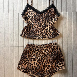 May include: A two-piece pyjama set featuring a camisole top and matching shorts with a brown and black leopard print pattern. The camisole has thin shoulder straps and black lace trim along the V-neckline. The shorts feature an elastic waistband.