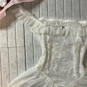 Może przedstawiać: Biała bielizna z koronkowymi detalami i falbankami. Odzież z odkrytymi ramionami ma delikatny kwiatowy wz&oacute;r koronki na gorsecie i zwiewną sp&oacute;dnicę. Miękki, przezroczysty materiał tworzy elegancki i romantyczny wygląd.