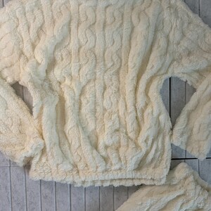 May include: Cream-colored, cable-knit style loungewear set. The top has long sleeves and a crew neck, with a cinched waist. The matching bottoms are also visible. The fabric appears soft and plush.
