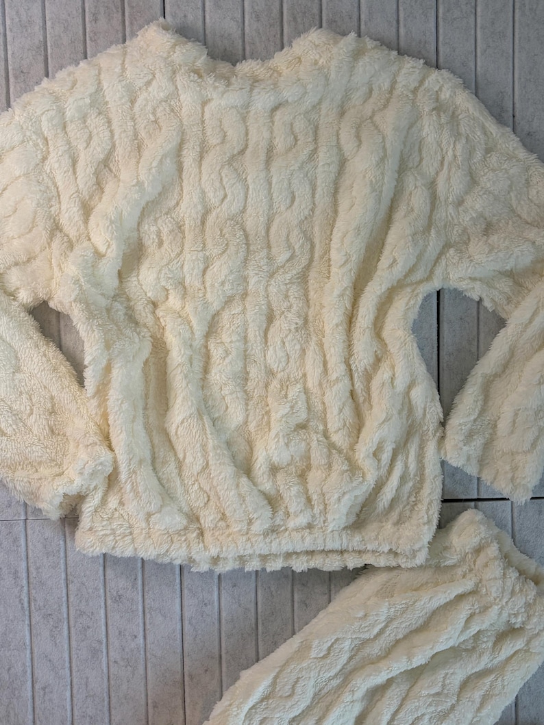 May include: Cream-colored, cable-knit style loungewear set. The top has long sleeves and a crew neck, with a cinched waist. The matching bottoms are also visible. The fabric appears soft and plush.