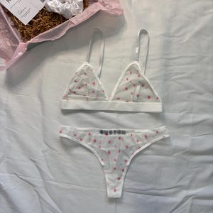 May include: White lingerie set with pink polka dots, including a triangle bralette and a thong. The thong features the word "CUSTOM" in silver letters. A white scrunchie and a gift box with a "Thank You" card are also visible.