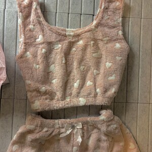 K&ouml;nnte beinhalten: Ein zweiteiliges Pyjama-Set aus weichem, pl&uuml;schigem Material. Das Set beinhaltet ein Crop-Top und Shorts, beide in Hellrosa mit einem wei&szlig;en Herzmuster. Die Shorts haben eine wei&szlig;e Schleife.