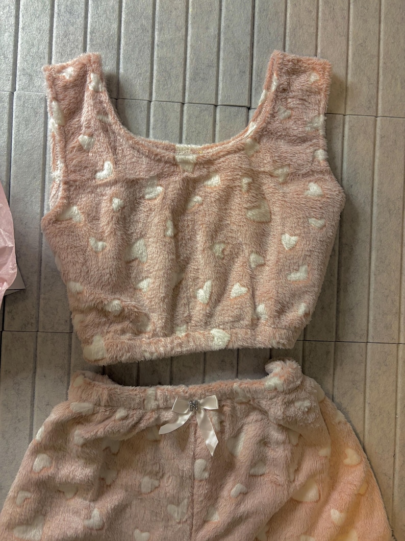 K&ouml;nnte beinhalten: Ein zweiteiliges Pyjama-Set aus weichem, pl&uuml;schigem Material. Das Set beinhaltet ein Crop-Top und Shorts, beide in Hellrosa mit einem wei&szlig;en Herzmuster. Die Shorts haben eine wei&szlig;e Schleife.