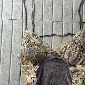 May include: A camisole featuring a dark mauve velvet bodice with cream-colored floral lace overlays on the cups and sides. The garment includes thin adjustable shoulder straps and a delicate string of small white faux pearls across the neckline.