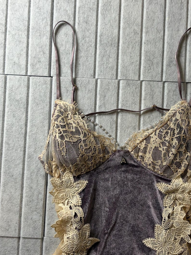 May include: A camisole featuring a dark mauve velvet bodice with cream-colored floral lace overlays on the cups and sides. The garment includes thin adjustable shoulder straps and a delicate string of small white faux pearls across the neckline.