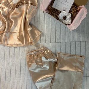 Op de afbeelding: Een champagnekleurige satijnen pyjamaset, bestaande uit een camisole top en broek, wordt getoond. Een geschenkdoos met een kaart met de tekst "Thank You" en een witte scrunchie is ook zichtbaar. De set is gerangschikt op een lichtgrijze ondergrond.