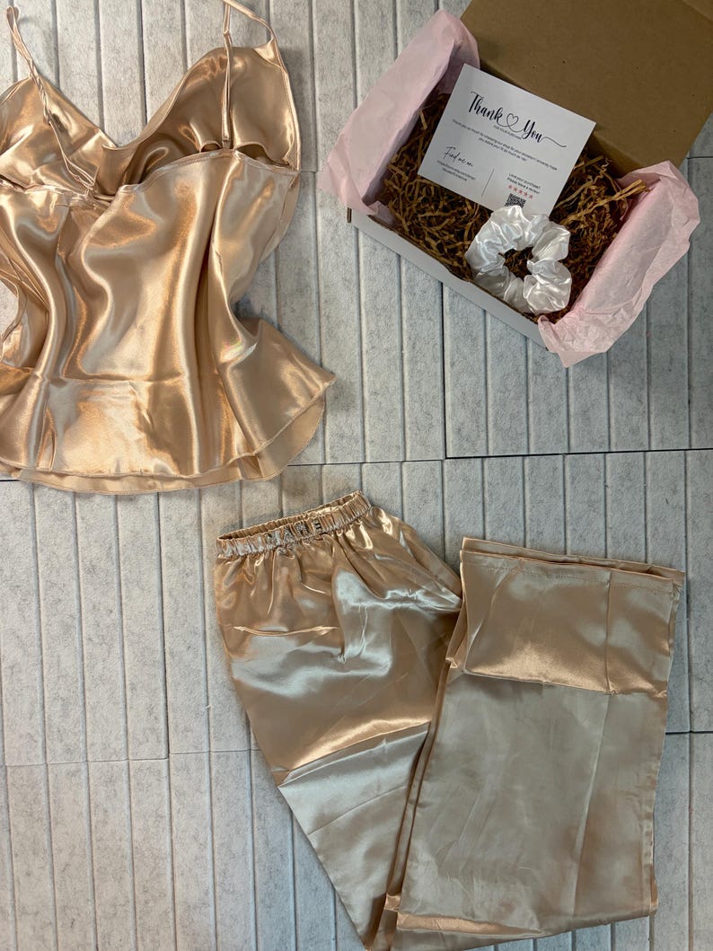 Op de afbeelding: Een champagnekleurige satijnen pyjamaset, bestaande uit een camisole top en broek, wordt getoond. Een geschenkdoos met een kaart met de tekst "Thank You" en een witte scrunchie is ook zichtbaar. De set is gerangschikt op een lichtgrijze ondergrond.