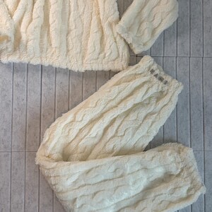 May include: Cream-colored, plush pajama set featuring a textured cable knit design. The set includes a long-sleeved top and matching pants. The pants have a small logo on the waistband. The fabric appears soft and comfortable.