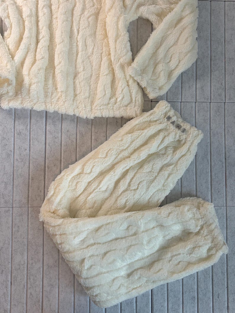 May include: Cream-colored, plush pajama set featuring a textured cable knit design. The set includes a long-sleeved top and matching pants. The pants have a small logo on the waistband. The fabric appears soft and comfortable.