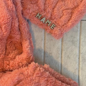 May include: A coral-colored, plush, fuzzy fabric with the word "NAME" spelled out in silver rhinestones. The fabric appears to be a soft, cozy material, possibly a blanket or a piece of clothing.