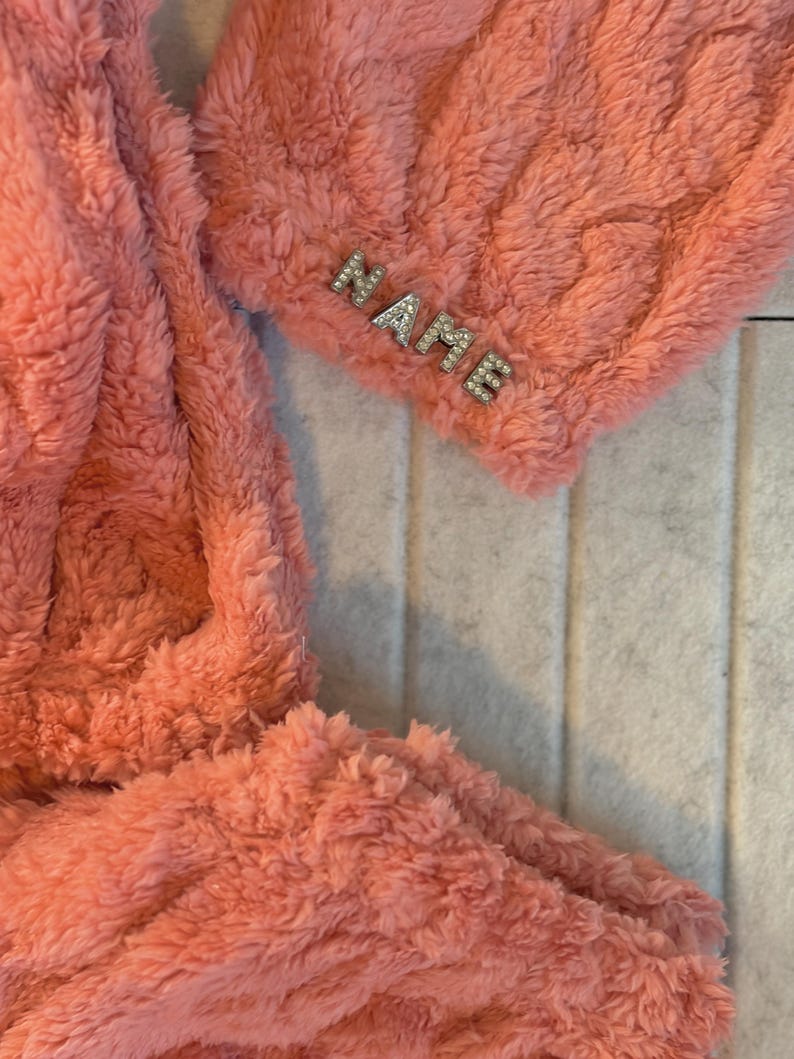May include: A coral-colored, plush, fuzzy fabric with the word "NAME" spelled out in silver rhinestones. The fabric appears to be a soft, cozy material, possibly a blanket or a piece of clothing.