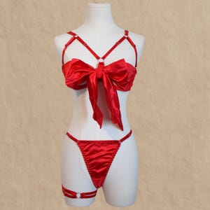 Puede incluir: Conjunto de lencería de satén rojo exhibido en un maniquí. El conjunto incluye un sujetador con un lazo grande, un tanga y una liga. El sujetador presenta finas correas y un detalle de anillo central.
