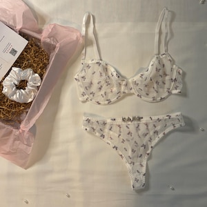May include: A white lingerie set with a butterfly pattern, including a bra and thong. A white scrunchie and a box with pink tissue paper are also visible. The lingerie set has a delicate, feminine design.