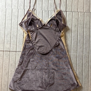 May include: A mauve velvet camisole featuring a deep scoop back, thin adjustable shoulder straps, and cream-colored lace trim along the neckline and sides. The garment has a soft, textured finish and a flared silhouette that falls to the hips.