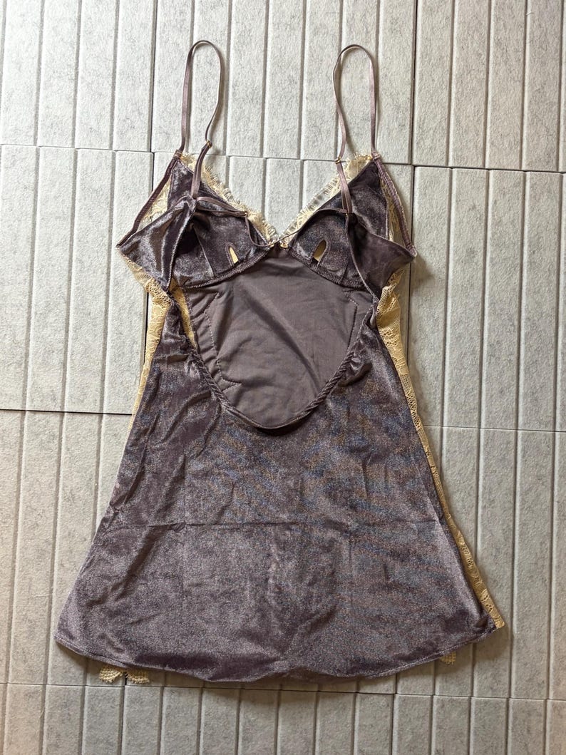 May include: A mauve velvet camisole featuring a deep scoop back, thin adjustable shoulder straps, and cream-colored lace trim along the neckline and sides. The garment has a soft, textured finish and a flared silhouette that falls to the hips.
