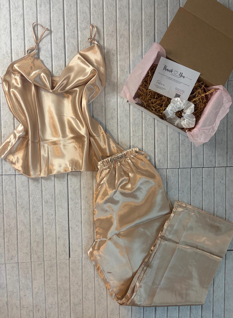 May include: A champagne-colored satin pajama set, including a camisole top with spaghetti straps and matching pants. A gift box with a "Thank You" card and a white scrunchie is also included. The set is displayed on a light gray tiled surface.