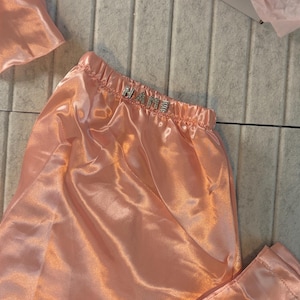 Op de afbeelding: Perzikroze satijnen pyjamaset met het woord "NAME" in zilveren strass-steentjes op de tailleband. De set bevat een broek en een bijpassende slaapmuts. De stof heeft een glanzende, gladde textuur.